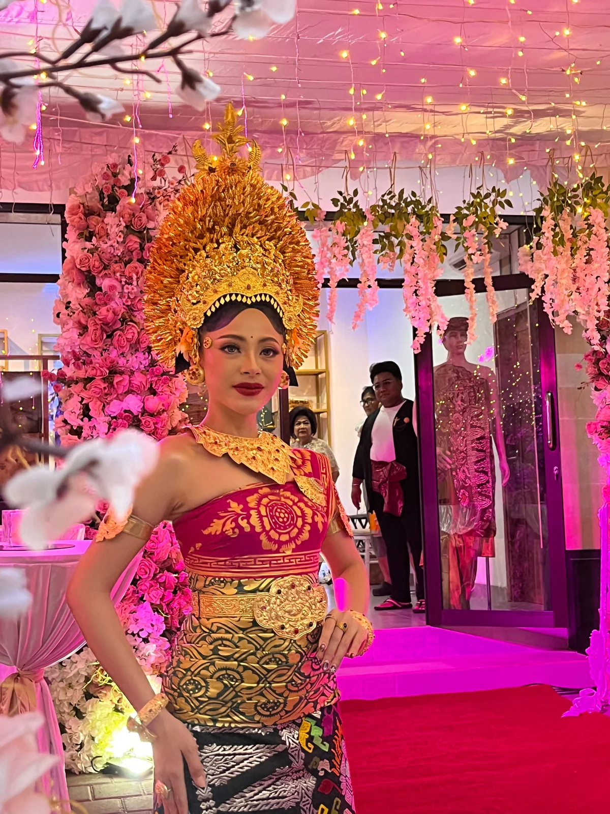 A bride wearing magenta and gold Balinese attire and an ornate gold headpiece beneath pink floral lighting