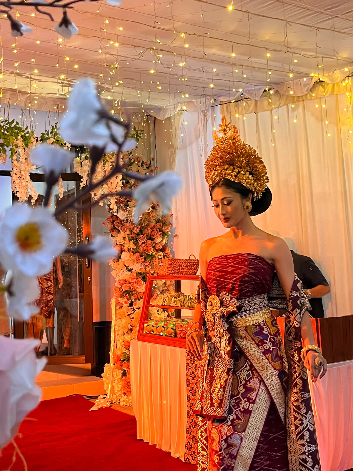 A bride in burgundy and gold ceremonial attire beside flowers and warm wedding lighting