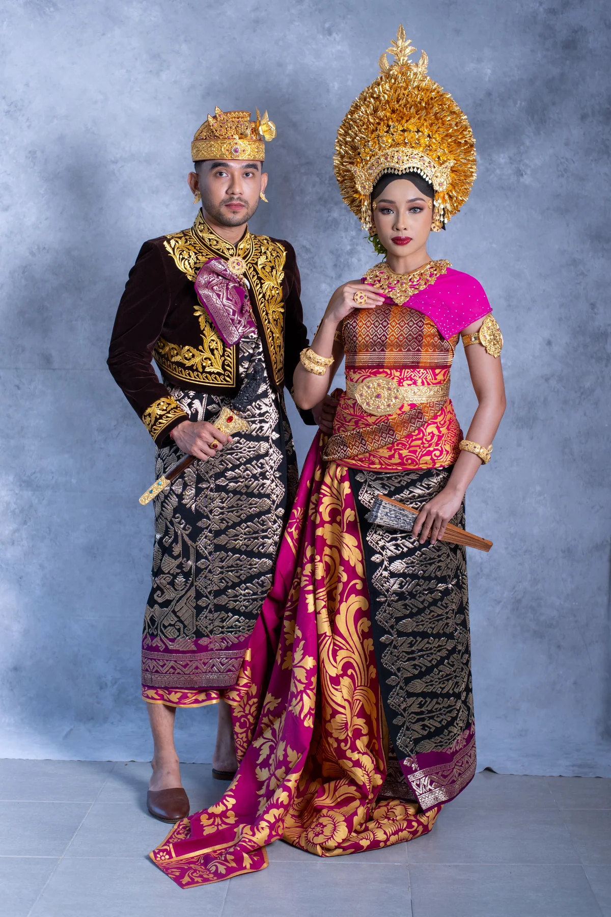 A bridal couple wearing coordinated magenta and gold Balinese ceremonial attire against a blue studio backdrop